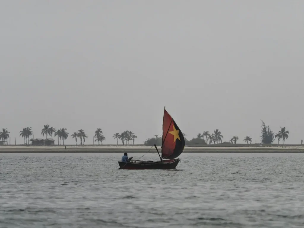 A fisherman sailing a small boat whose sail carries the red, black and yellow star of the Angolan flag, off a palm-lined shore in Angola