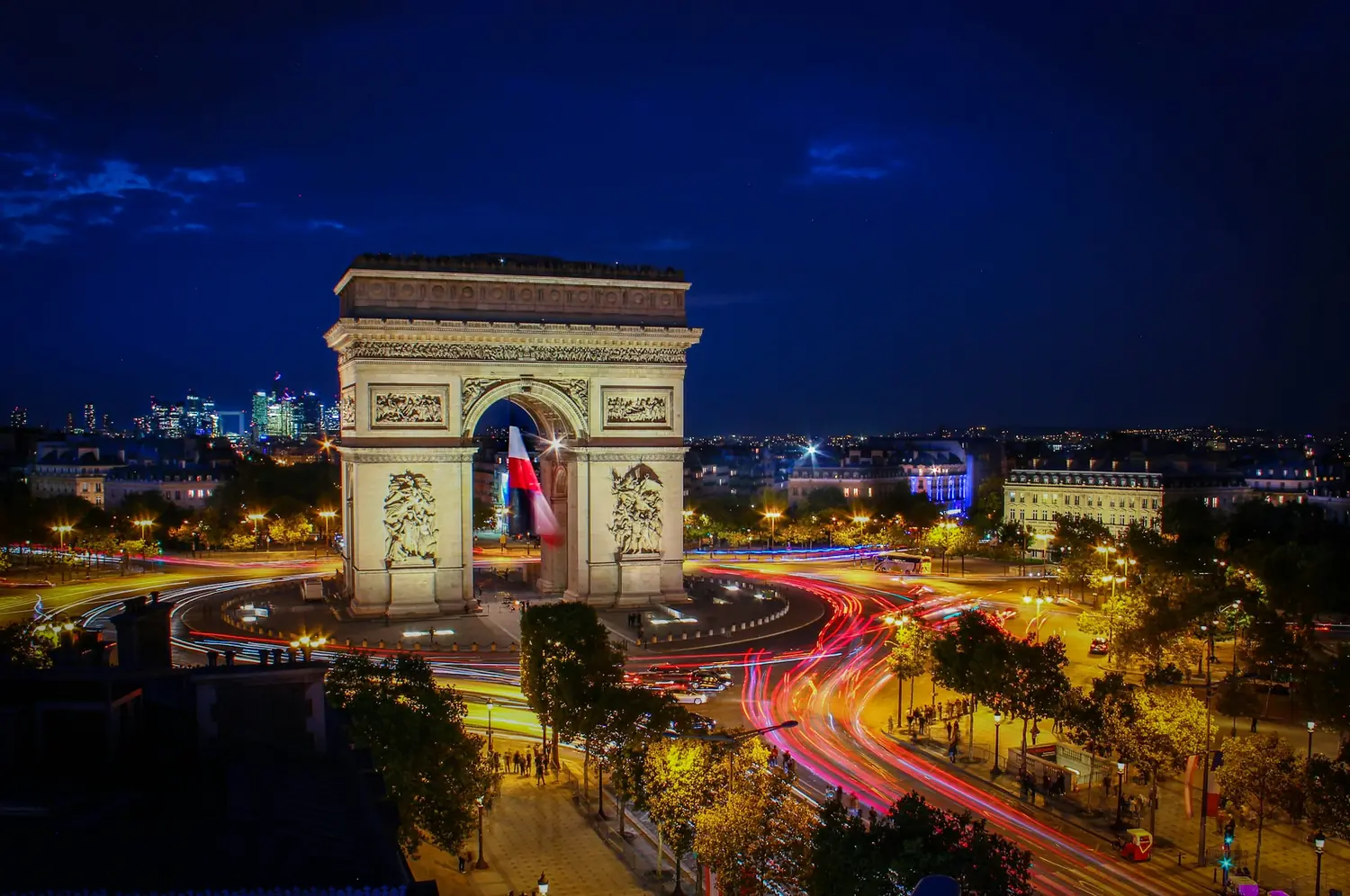 The Arc de Triomphe lit up at night with traffic trails around the Place Charles de Gaulle and La Defense in the distance, Paris, France