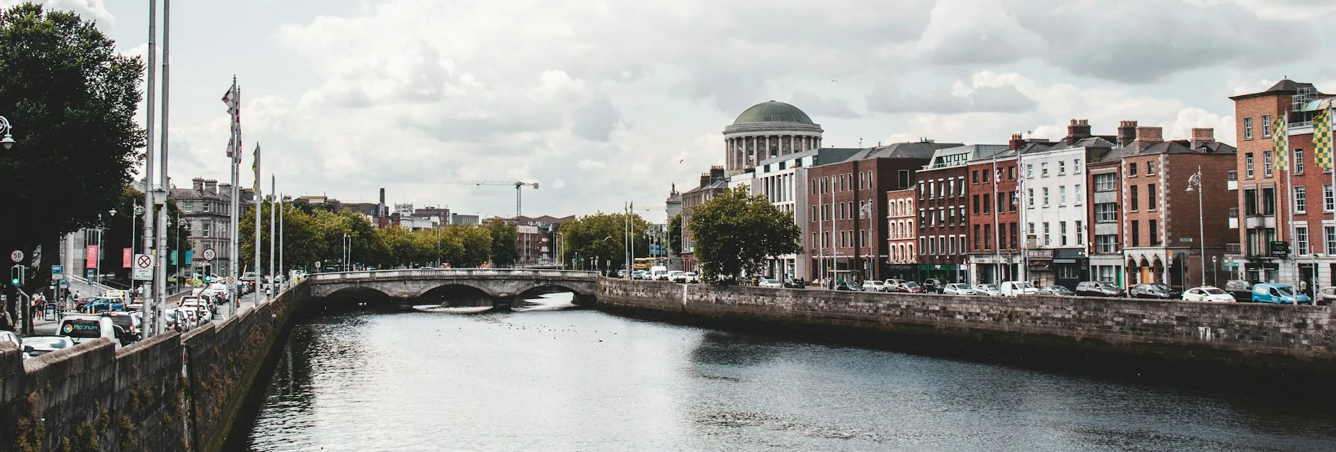 The River Liffey in Dublin seen from a bridge, with the quays on both banks lined by offices, shops and parked cars, the dome of the Four Courts on the north side and a construction crane behind, Dublin, Ireland