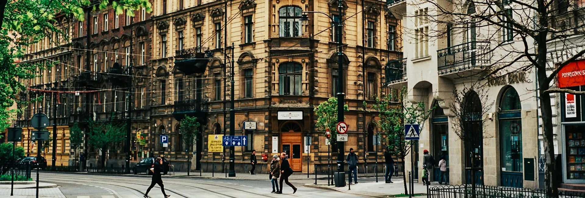 People crossing a tram street lined with 19th-century tenement buildings and shopfronts in central Kraków, Poland