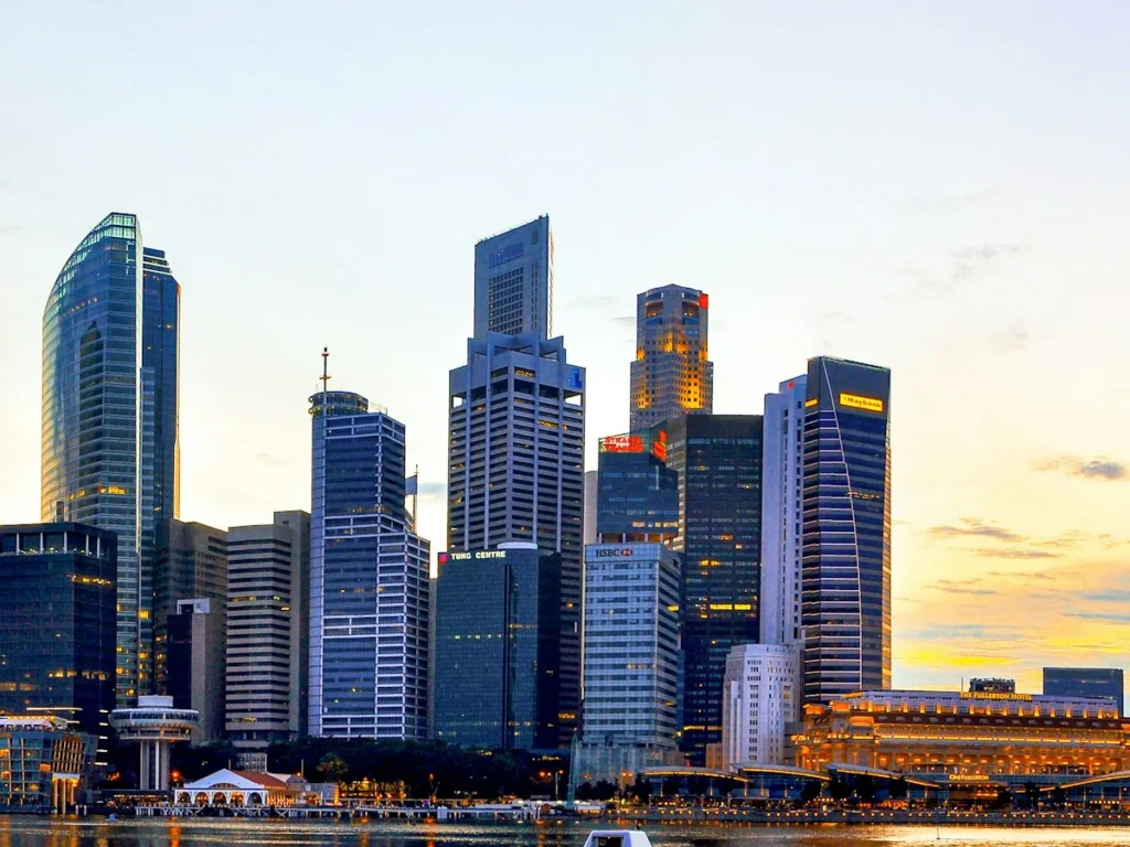 Office towers in the Singapore central business district at dusk, with the Fullerton Hotel and the Marina Bay waterfront lit below and the sunset behind, Singapore