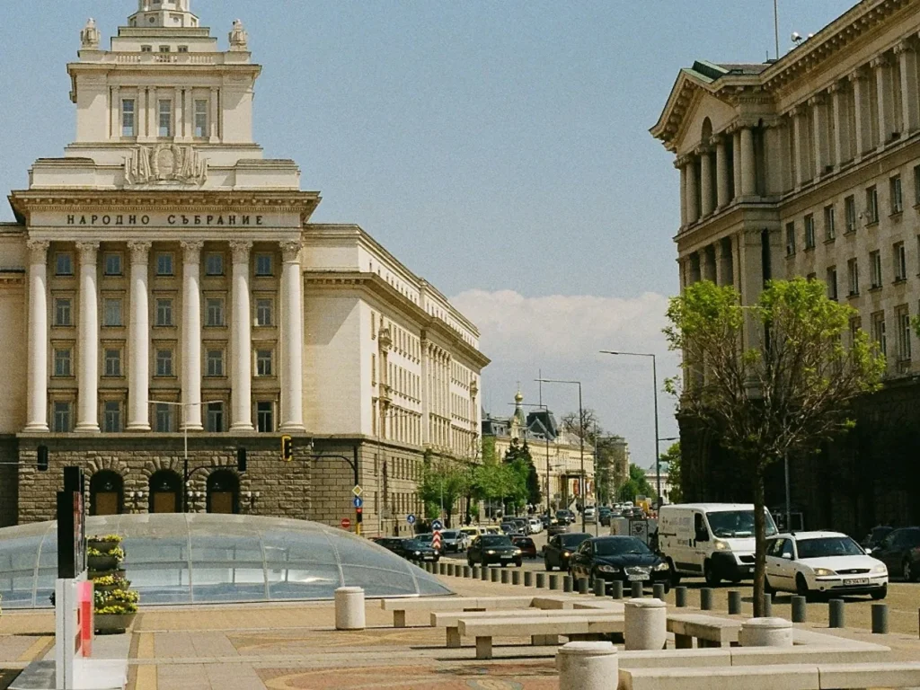 Traffic on the boulevard through central Sofia at midday, with the stone facade and columns of the Largo on one side, parked cars and street trees on the other, Sofia, Bulgaria