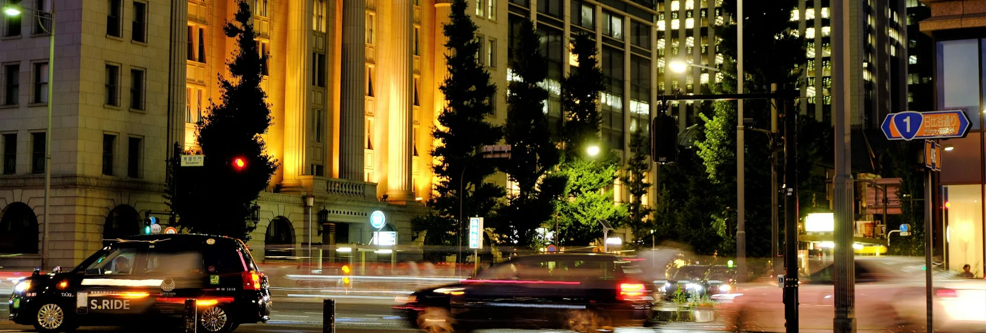 Marunouchi at night on Hibiya-dori, with the lit colonnade of the Meiji Seimeikan, office towers with their lights on behind it and a taxi and traffic passing, Tokyo, Japan