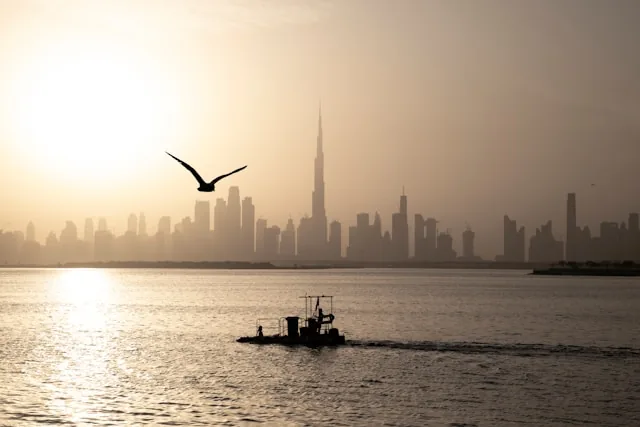 The Dubai skyline in silhouette at dawn seen across the water, with the Burj Khalifa at the centre and a working boat in the foreground, UAE