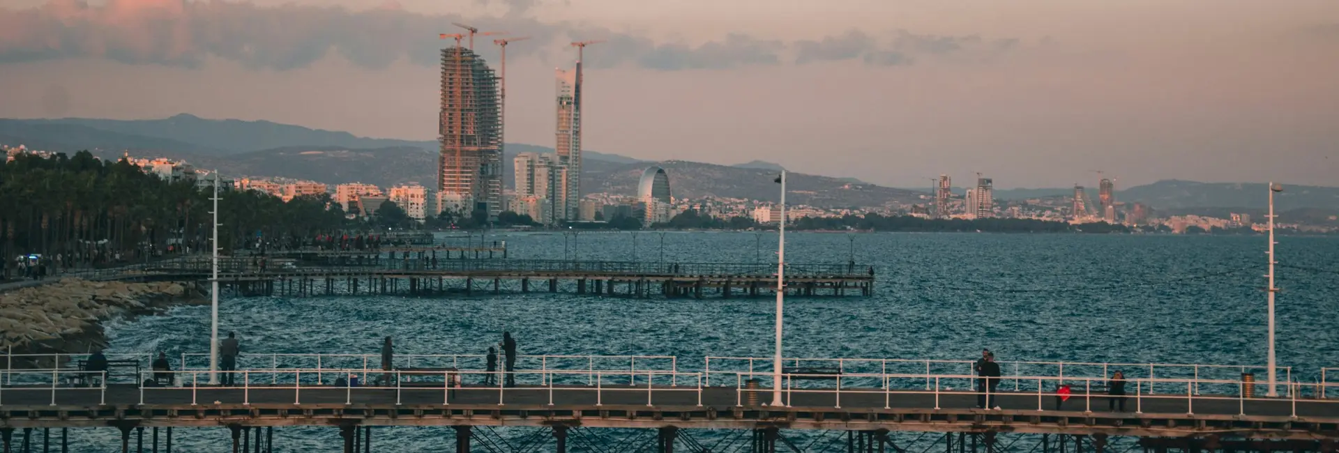 People fishing from a pier on the Limassol seafront at sunset, with high-rise towers along the coast, Cyprus